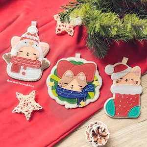 May include: Felt Christmas ornaments featuring cat designs. One ornament is shaped like a snowman, another has a wreath, and the third is a cat in a stocking. Each cat wears a scarf or hat. The ornaments are on a red surface.