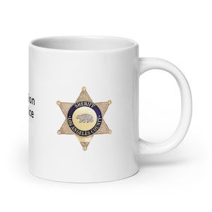 May include: White ceramic mug with a gold and blue Los Angeles County Sheriff's star badge printed on the front. The badge features a bear and the words "Sheriff Los Angeles County".