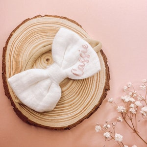 May include: A white fabric bow headband with the name "Cassie" embroidered in pink on a wooden background.