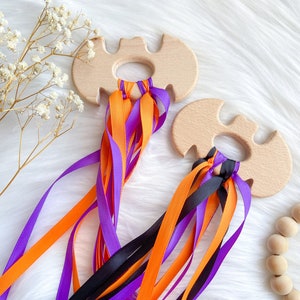 Puede incluir: Dos juguetes de madera con forma de murci&eacute;lago con bucles para los dedos. Cada juguete tiene m&uacute;ltiples cintas de color naranja, morado y negro.