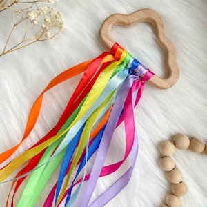 K&ouml;nnte beinhalten: Ein Holz-Bei&szlig;ring in Wolkenform mit daran befestigten regenbogenfarbenen Satinb&auml;ndern. Der Bei&szlig;ring ist aus Naturholz gefertigt und f&uuml;r Babys sicher.