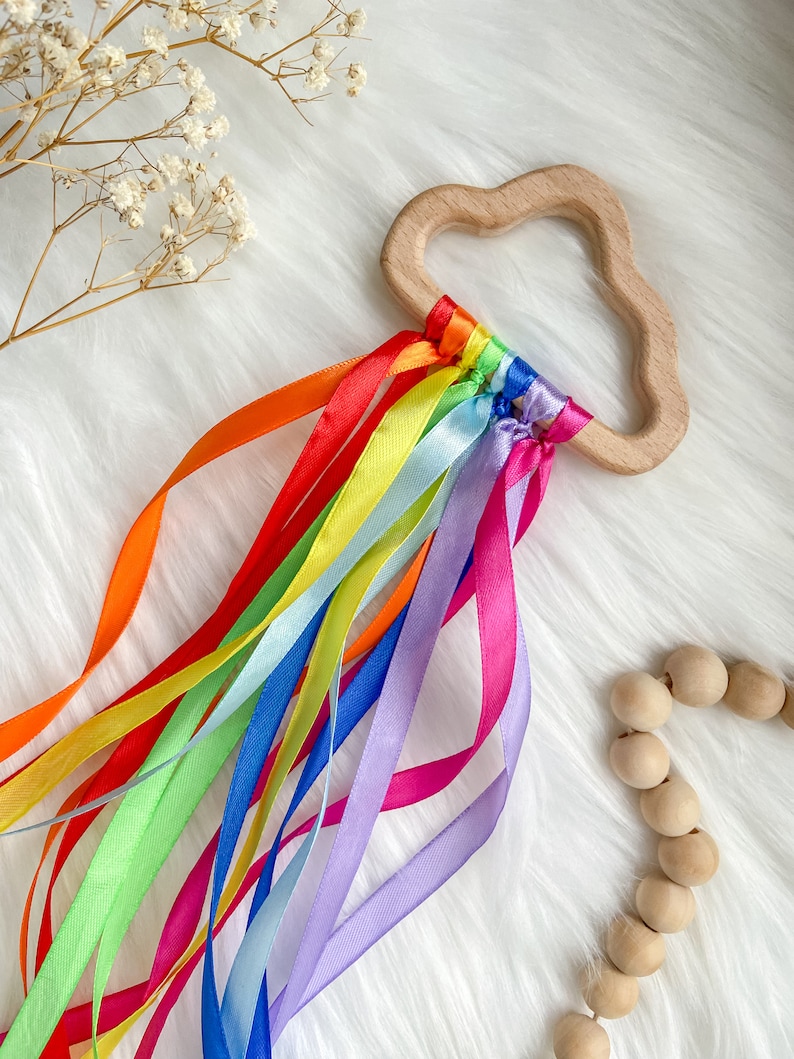 K&ouml;nnte beinhalten: Ein Holz-Bei&szlig;ring in Wolkenform mit daran befestigten regenbogenfarbenen Satinb&auml;ndern. Der Bei&szlig;ring ist aus Naturholz gefertigt und f&uuml;r Babys sicher.