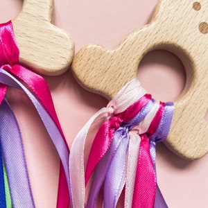 K&ouml;nnte beinhalten: Zwei Holzringe mit bunten B&auml;ndern befestigt. Die Ringe sind aus Naturholz und haben eine glatte Oberfl&auml;che. Die B&auml;nder sind rosa, lila und wei&szlig;.