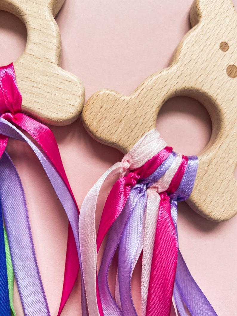 K&ouml;nnte beinhalten: Zwei Holzringe mit bunten B&auml;ndern befestigt. Die Ringe sind aus Naturholz und haben eine glatte Oberfl&auml;che. Die B&auml;nder sind rosa, lila und wei&szlig;.