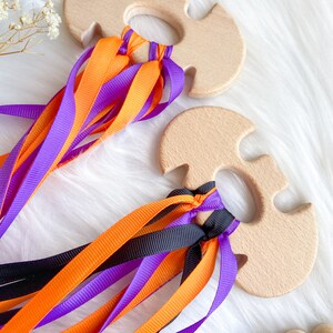 Puede incluir: Dos juguetes de madera con forma de murci&eacute;lago con cintas de colores naranja, morado y negro. Los juguetes est&aacute;n hechos de madera natural y tienen un acabado liso.