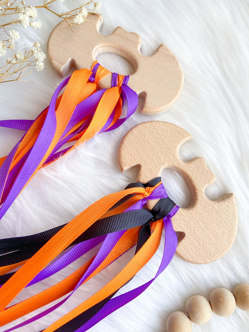 Puede incluir: Dos juguetes de madera con forma de murci&eacute;lago con cintas de colores naranja, morado y negro. Los juguetes est&aacute;n hechos de madera natural y tienen un acabado liso.