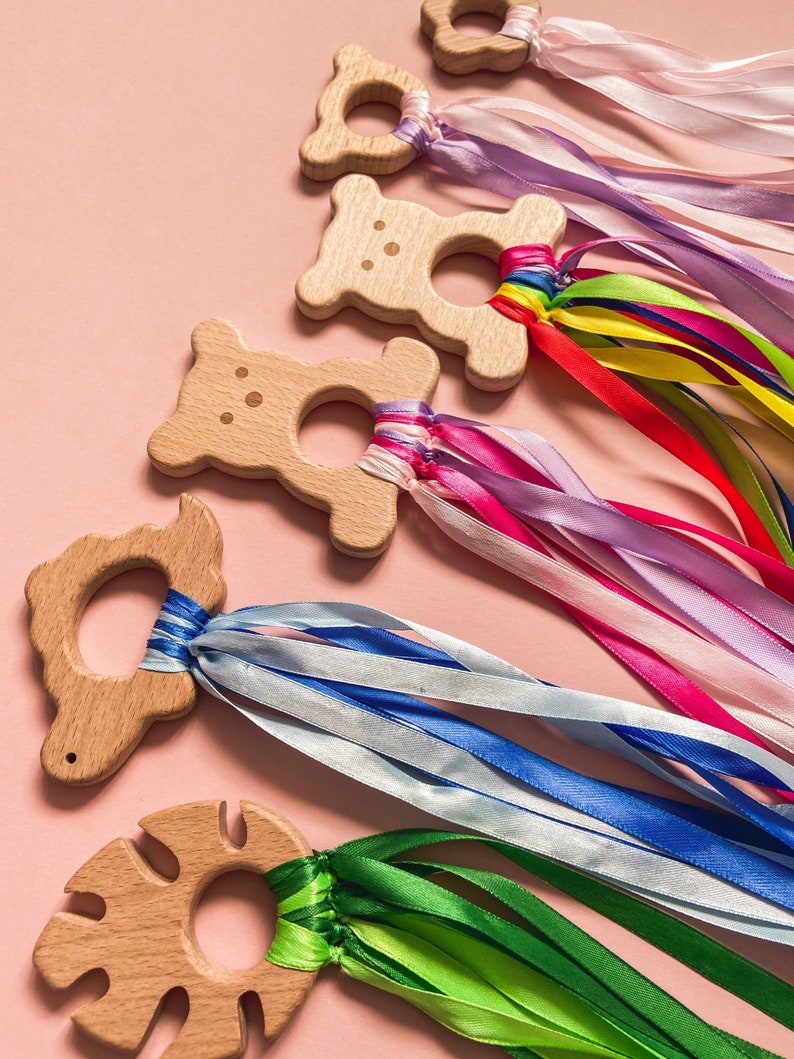 K&ouml;nnte beinhalten: F&uuml;nf Holz-Bei&szlig;ringe mit bunten Schleifenb&auml;ndern. Die Bei&szlig;ringe sind in Form von B&auml;ren, Schildkr&ouml;ten und Bl&auml;ttern. Die B&auml;nder sind in den Farben Rosa, Blau, Gr&uuml;n und Gelb.