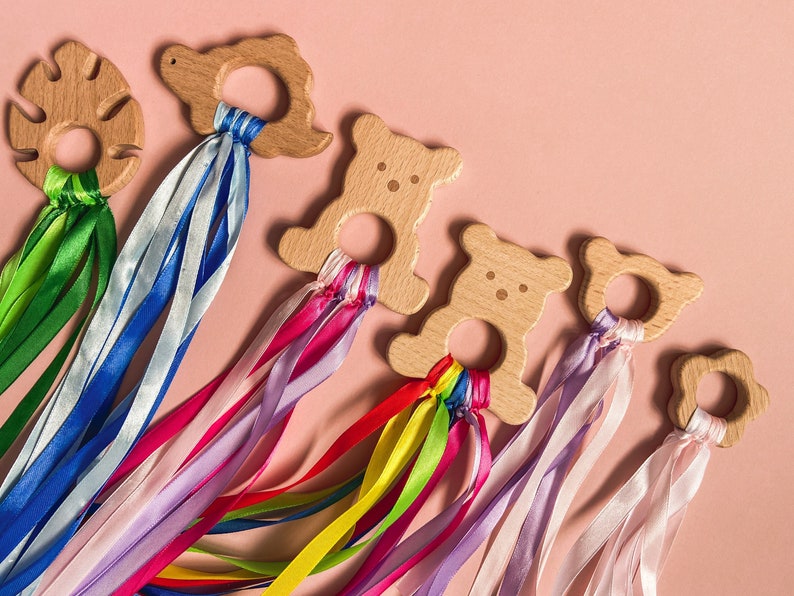 K&ouml;nnte beinhalten: F&uuml;nf Holz-Bei&szlig;ringe mit bunten B&auml;ndern befestigt. Die Bei&szlig;ringe sind in Form eines Blattes, eines Dinosauriers, eines B&auml;ren, eines B&auml;ren und einer Blume. Die B&auml;nder sind oben an jedem Bei&szlig;ring zusammengebunden.