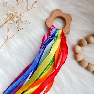 K&ouml;nnte beinhalten: Ein sensorisches Holzspielzeug in Regenbogenfarben mit einem blumenf&ouml;rmigen Ring und bunten B&auml;ndern. Das Spielzeug ist perfekt f&uuml;r Babys und Kleinkinder, um ihre Sinne zu erforschen.