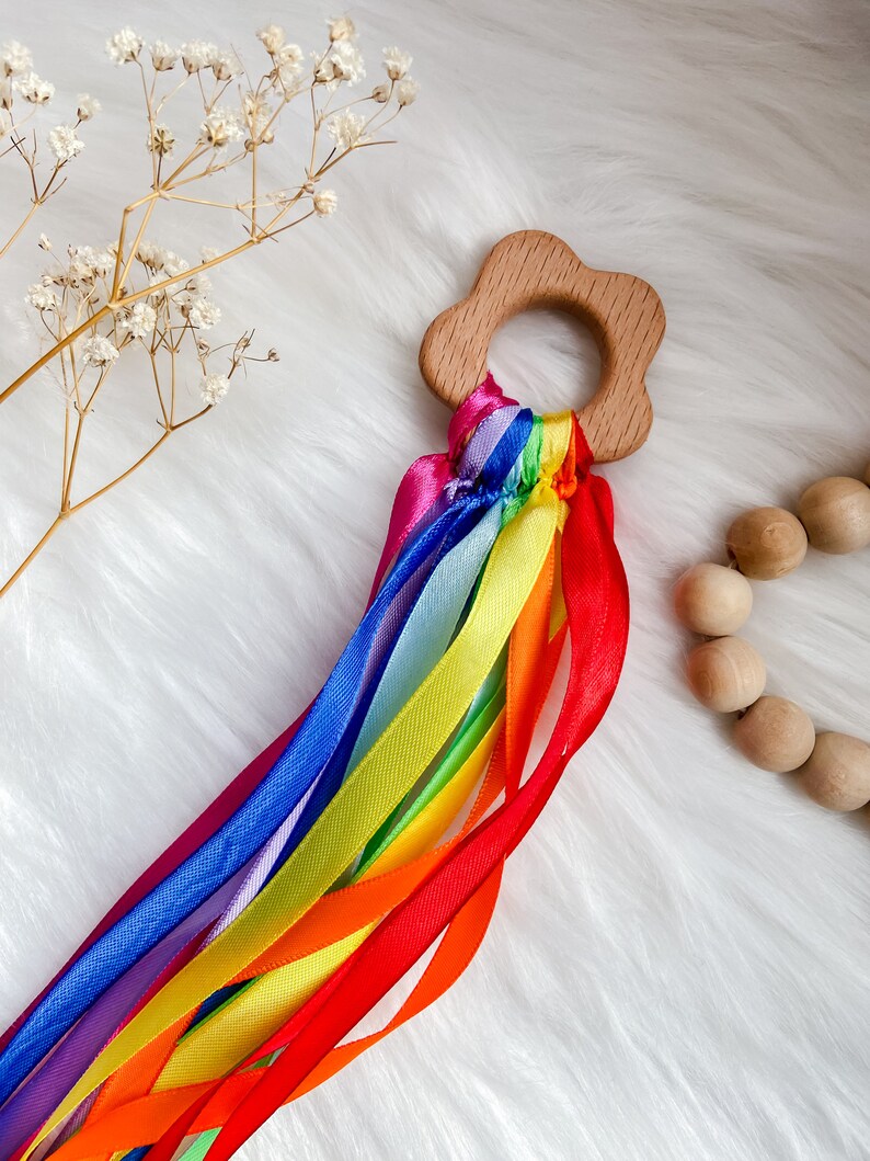 K&ouml;nnte beinhalten: Ein sensorisches Holzspielzeug in Regenbogenfarben mit einem blumenf&ouml;rmigen Ring und bunten B&auml;ndern. Das Spielzeug ist perfekt f&uuml;r Babys und Kleinkinder, um ihre Sinne zu erforschen.