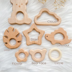 K&ouml;nnte beinhalten: Satz von zehn Holz-Bei&szlig;ringen in verschiedenen Formen, darunter ein Teddyb&auml;r, eine Wolke, ein Stern, ein Blatt, eine Blume, ein B&auml;renkopf, ein Dinosaurier und ein Kreis. Die Bei&szlig;ringe sind aus Naturholz gefertigt und perfekt f&uuml;r Babys zum Kauen.