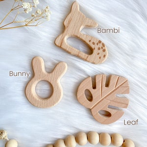 K&ouml;nnte beinhalten: Vier Holz-Bei&szlig;ringe in Form eines Rehs, eines Hasen, eines Blattes und einer Schnur aus Holzperlen. Die Bei&szlig;ringe sind in nat&uuml;rlicher Holzfarbe und haben eine glatte Oberfl&auml;che. Der Reh-Bei&szlig;ring hat das Wort "Bambi" daneben geschrieben, der Hasen-Bei&szlig;ring hat das Wort "Bunny" daneben geschrieben und der Blatt-Bei&szlig;ring hat das Wort "Leaf" daneben geschrieben.