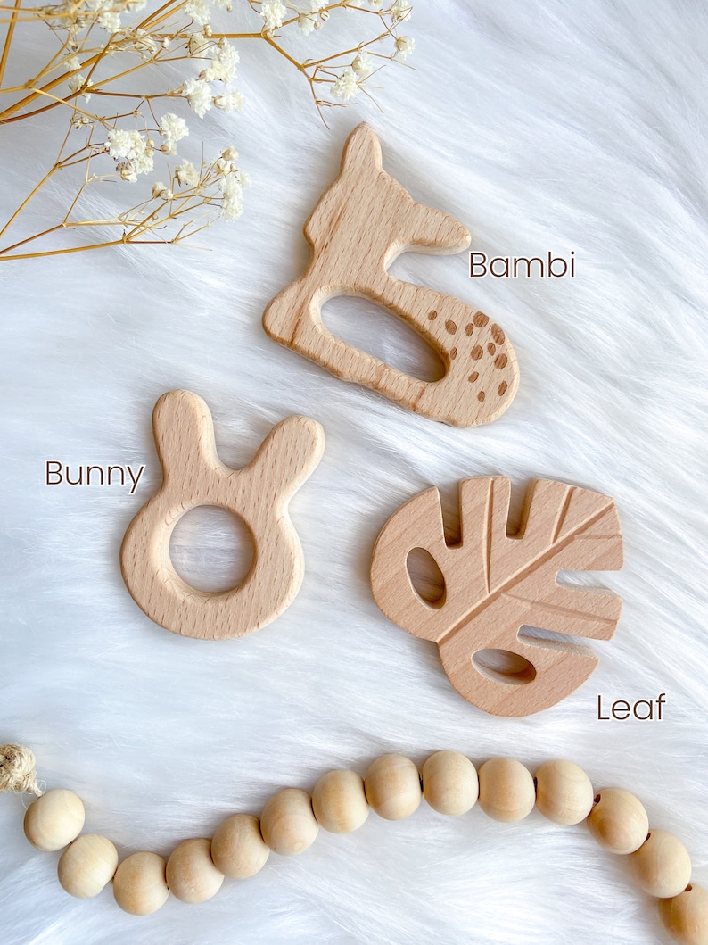 K&ouml;nnte beinhalten: Vier Holz-Bei&szlig;ringe in Form eines Rehs, eines Hasen, eines Blattes und einer Schnur aus Holzperlen. Die Bei&szlig;ringe sind in nat&uuml;rlicher Holzfarbe und haben eine glatte Oberfl&auml;che. Der Reh-Bei&szlig;ring hat das Wort "Bambi" daneben geschrieben, der Hasen-Bei&szlig;ring hat das Wort "Bunny" daneben geschrieben und der Blatt-Bei&szlig;ring hat das Wort "Leaf" daneben geschrieben.