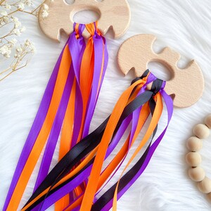 Puede incluir: Dos mordedores de madera en forma de murci&eacute;lago con cintas de color naranja, morado y negro atadas. Los mordedores est&aacute;n hechos de madera natural y tienen un acabado liso.