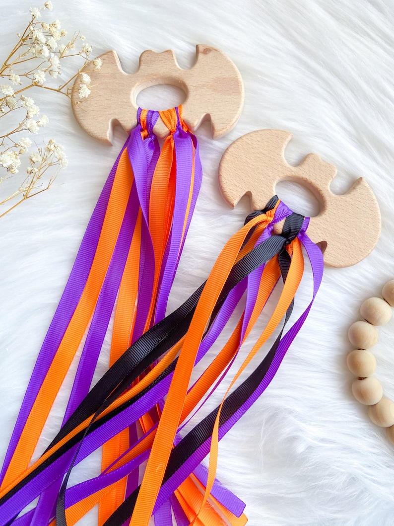Puede incluir: Dos mordedores de madera en forma de murci&eacute;lago con cintas de color naranja, morado y negro atadas. Los mordedores est&aacute;n hechos de madera natural y tienen un acabado liso.