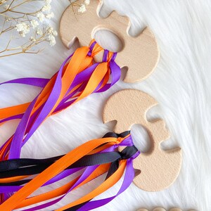 Puede incluir: Dos mordedores de madera con forma de murci&eacute;lago con cintas de color naranja, morado y negro atadas. Los mordedores est&aacute;n hechos de madera natural y tienen un acabado liso.