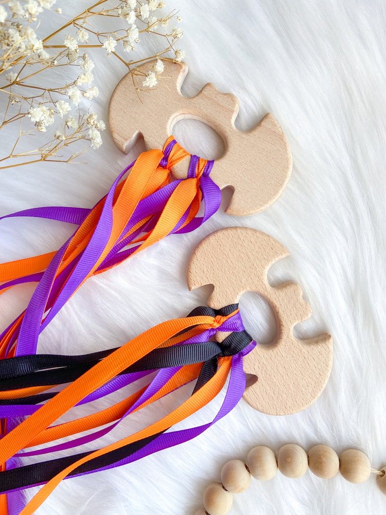 Puede incluir: Dos mordedores de madera con forma de murci&eacute;lago con cintas de color naranja, morado y negro atadas. Los mordedores est&aacute;n hechos de madera natural y tienen un acabado liso.