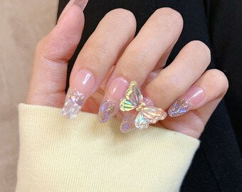 May include: Close-up of a hand with long, clear, oval-shaped fake nails decorated with iridescent butterfly charms and glitter.
