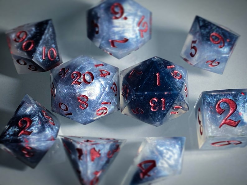 May include: A set of ten-sided dice with a galaxy-like pattern and red numbers. The dice are made of a translucent material and have a sparkly finish.