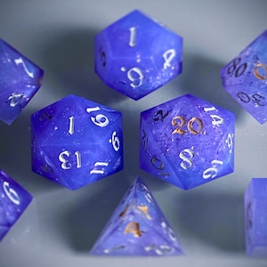 May include: A set of eight purple and blue dice with white and gold numbers. The dice are made of a translucent material and have a speckled appearance. The numbers on the dice are 1, 2, 3, 4, 5, 6, 8, 9, 13, 14, 16, 20, and 61.