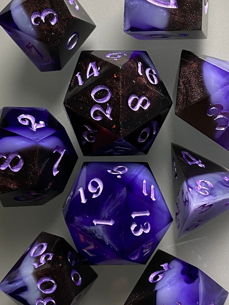May include: A set of seven purple and black dice with white numbers. The dice are made of a translucent material with a swirling pattern.