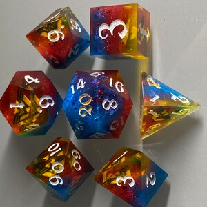May include: Set of seven polyhedral dice with a rainbow glitter finish. The dice are numbered 1-6, 1-20, 4-sided, 6-sided, 8-sided, 10-sided, and 12-sided. The dice are a mix of red, yellow, blue, and green.