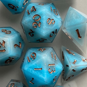 May include: A set of eight blue and white dice with gold numbers. The dice are made of a translucent material with a glittery finish. The numbers on the dice are 1, 2, 3, 4, 5, 6, 8, 10, 12, 14, 16, 18, 19, 20.