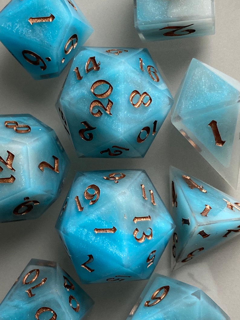 May include: A set of eight blue and white dice with gold numbers. The dice are made of a translucent material with a glittery finish. The numbers on the dice are 1, 2, 3, 4, 5, 6, 8, 10, 12, 14, 16, 18, 19, 20.