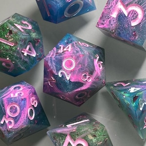 May include: Set of six iridescent dice with pink and blue swirls and glitter. The dice are numbered 1 through 6 with white numbers and pink outlines.