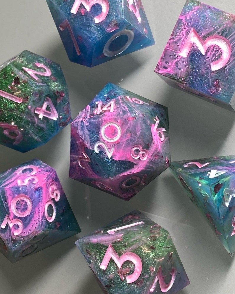 May include: Set of six iridescent dice with pink and blue swirls and glitter. The dice are numbered 1 through 6 with white numbers and pink outlines.