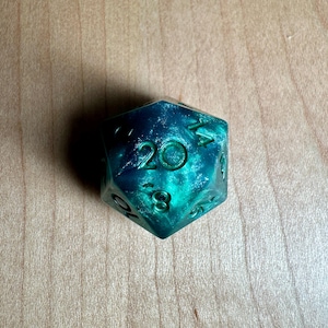 Peut inclure: Un dé à 20 faces de couleur vert turquoise avec des chiffres noirs. Le dé a une finition scintillante.