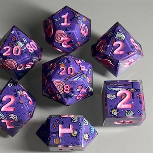 May include: Set of seven purple and pink dice with a variety of whimsical designs. The dice are numbered 1 through 20.