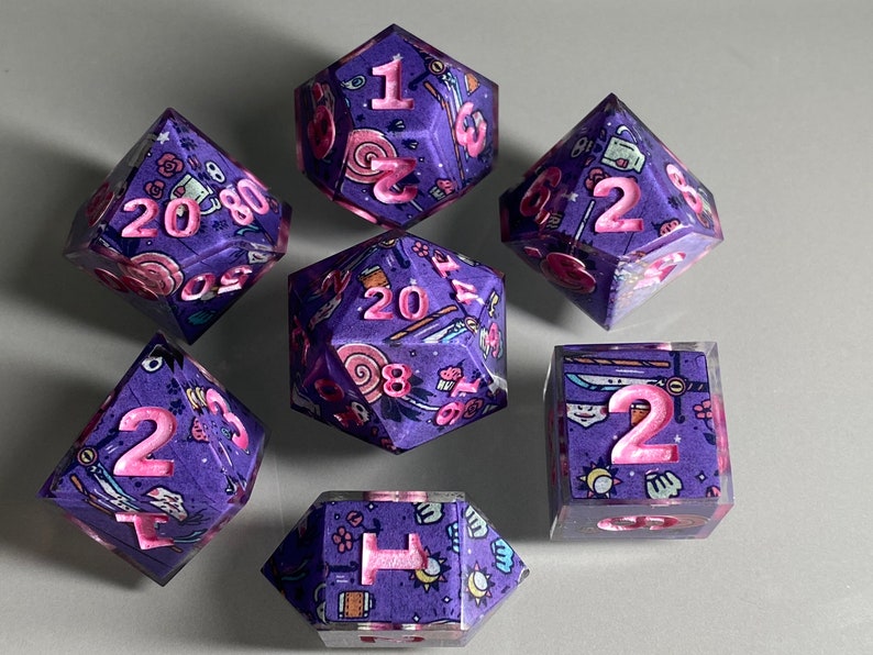 May include: Set of seven purple and pink dice with a variety of whimsical designs. The dice are numbered 1 through 20.
