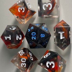 May include: Set of seven polyhedral dice with black, white, and orange swirl patterns. The dice are made of clear resin and have white numbers. The dice are for tabletop role-playing games.