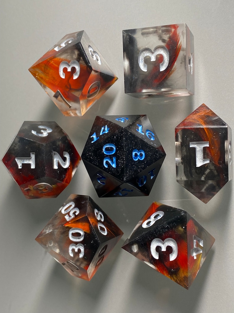May include: Set of seven polyhedral dice with black, white, and orange swirl patterns. The dice are made of clear resin and have white numbers. The dice are for tabletop role-playing games.