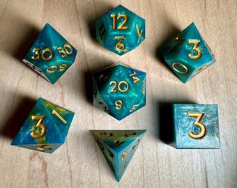 May include: A set of seven teal and gold polyhedral dice with a glittery finish. The dice are numbered 3, 4, 6, 8, 12, 20, and 30.