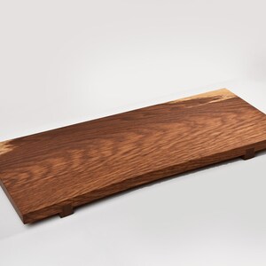 Peut inclure: Planche de service rectangulaire en bois avec un fini grain de bois naturel. La planche est principalement de couleur marron foncé, avec une teinte de bois plus claire et naturelle à une extrémité. Elle a de petits pieds en dessous.
