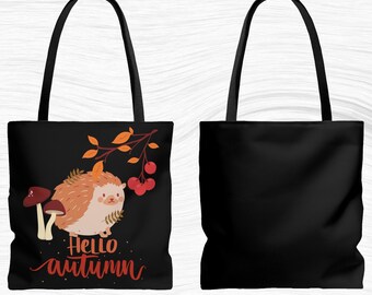 May include: Black tote bag featuring a cartoon hedgehog with autumn leaves, mushrooms, and berries. The words "Hello Autumn" are written in a cursive font. The bag has black handles and a solid black back.