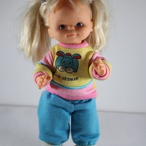 May include: A blonde doll wearing a pink and yellow sweater with a blue bunny graphic and blue pants. The doll is on roller skates with yellow wheels.