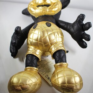 May include: A black and gold plush toy of Mickey Mouse. The toy is wearing a gold quilted outfit with black accents. The toy is standing on a white surface.
