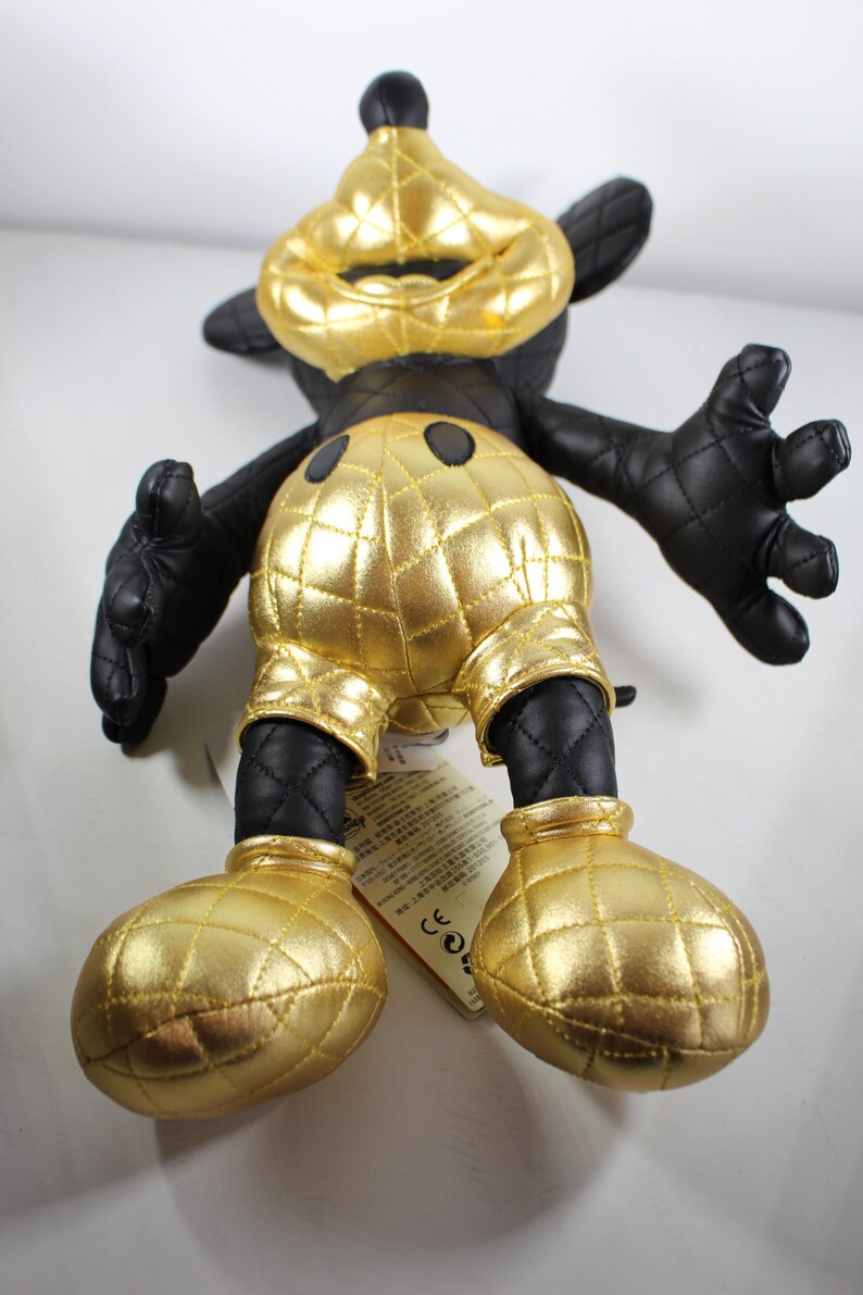 May include: A black and gold plush toy of Mickey Mouse. The toy is wearing a gold quilted outfit with black accents. The toy is standing on a white surface.