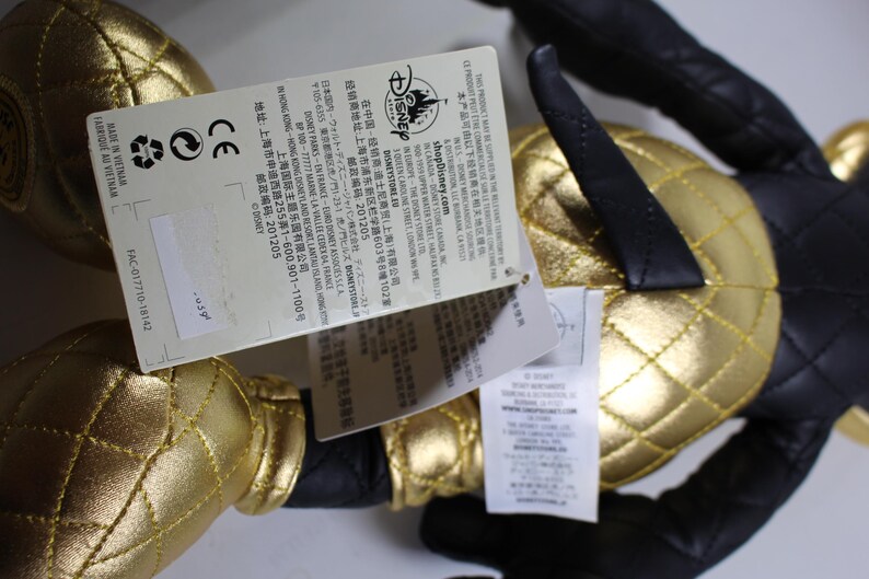 May include: A black and gold plush toy with a quilted design. The toy has a tag attached with the Disney logo and text in Japanese and English. The text on the tag includes the website shopDisney.com and the address of the Disney Store in Shanghai, China.