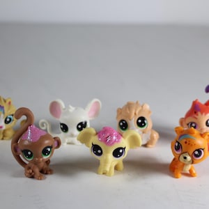 May include: A collection of seven small plastic animal figurines. The figurines are in various colors, including white, brown, orange, pink, and yellow. They have large eyes and are designed to look like cartoon animals.