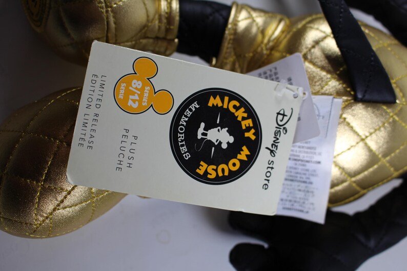 May include: Gold Mickey Mouse plush toy with a black quilted fabric. The tag reads "Disney Store Mickey Mouse Memories Plush Limited Release Edition Limit&eacute;e 8/12 Series".