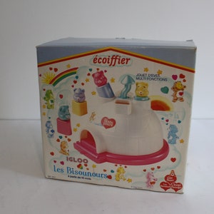 Peut inclure: Un jeu de construction en forme d'igloo pour les Bisounours. La boîte est blanche et rose avec un arc-en-ciel et le texte "écoinfier Igloo les Bisounours à partir de 18 mois".