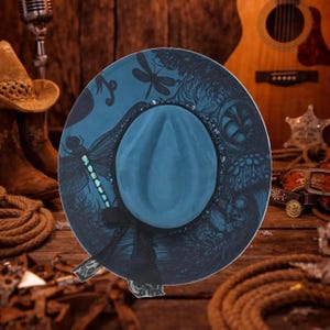 Può includere: Un cappello a tesa larga color verde acqua con una stampa scura e dettagliata di libellule ed elementi botanici sulla corona e sulla tesa. Il cappello è decorato con un nastro nero annodato a fiocco e presenta una piega centrale.