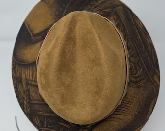 May include: A brown suede fedora hat with a pinched crown and a wide brim. The brim features a dark brown printed pattern depicting wheat stalks and western-style boots. A thin leather band encircles the base of the crown, accented with small silver-toned metal studs.
