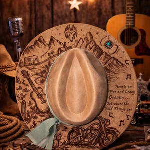 May include: A tan felt hat with a light green ribbon band. The brim features detailed pyrography illustrations of mountains, a guitar, a motorcycle, and a camper. The text reads: Hearts on Fire and Crazy Dreams... Out where the Wild Things are.