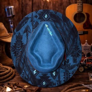 May include: A blue felt hat featuring a dark black print of feathers, arrows, and geometric patterns. The crown and brim are accented with small, light blue turquoise-coloured stones arranged in a vertical line and scattered around the base.