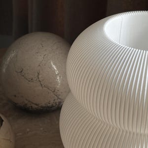 May include: A white vase with a rounded, stacked shape and a vertical ribbed texture. Beside it sits a spherical object with a mottled, stone-like surface featuring dark grey cracks and a small, circular candle holder with a metallic rim.